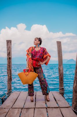 Editorial view on a wooden dock by turquoise water featuring a golden-yellow handwoven recycled plastic resort tote bag with red handles made in Guatemala