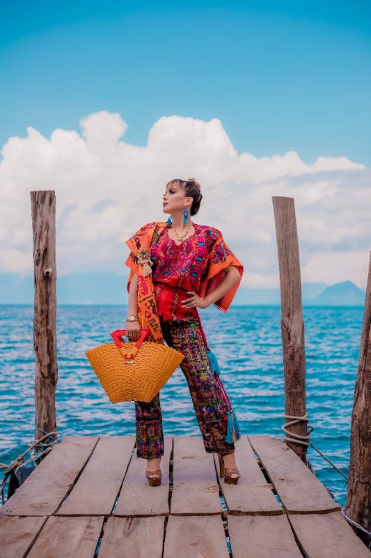 Editorial view on a wooden dock by turquoise water featuring a golden-yellow handwoven recycled plastic resort tote bag with red handles made in Guatemala